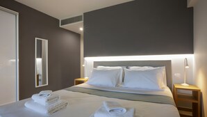 Ein Hotelzimmer mit einem ordentlich bezogenen Bett, einer Nachttischlampe, einem Spiegel und einem Wandlüfter.