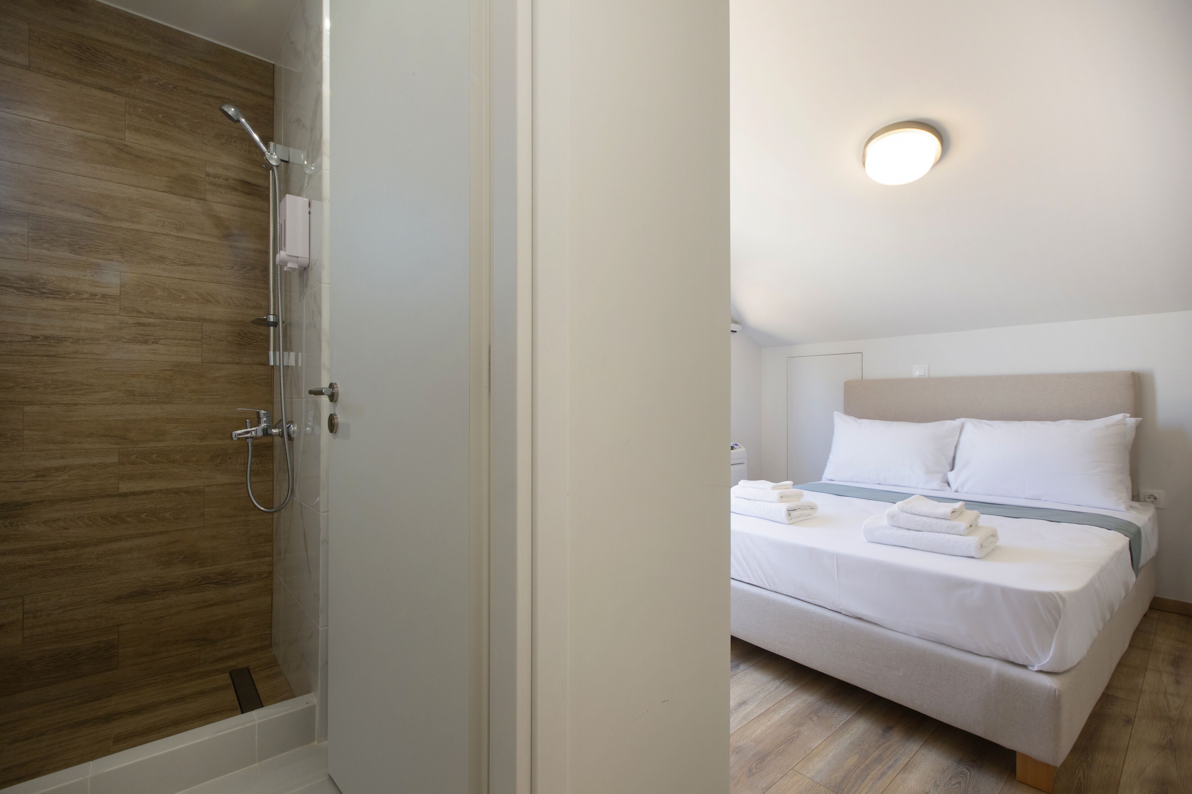 A hotel room with a wooden floor, a bed with white bedding, and a shower area with a glass door.