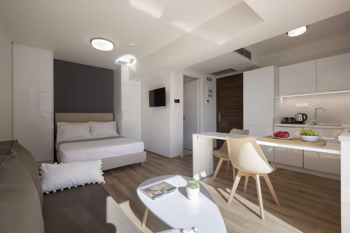 Une chambre moderne et bien éclairée, avec un lit, une petite table sur laquelle se trouve un magazine et une plante, ainsi qu’un coin repas séparé comprenant une chaise et un comptoir pour prendre le petit-déjeuner.