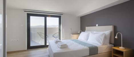 A modern hotel room with a large bed, a bedside table with a lamp, a sliding glass door leading to a balcony, and a view of the cityscape.