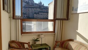 Una habitación con dos sillones de madera y una mesita con libros y una lámpara, una ventana que ofrece vistas a un edificio histórico, y un suelo de baldosas.