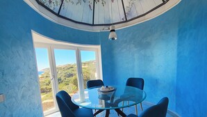 Un comedor con una mesa de vidrio y cuatro sillas, ventanas amplias con vistas a zonas verdes y colinas, y un techo con diseño floral.