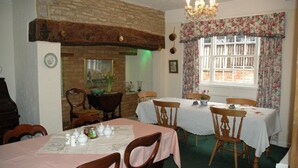 A dining area with brick walls, wooden tables, and chairs. There is a chandelier, a window with floral curtains, and a fireplace with a vase of flowers. - Pear Tree Farm B&B (Kettering)