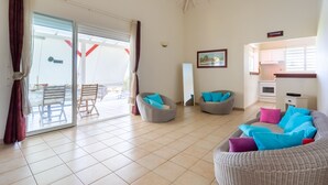 A spacious living area with a sliding glass door leading to an outdoor patio, wicker furniture, and a kitchenette in the background.