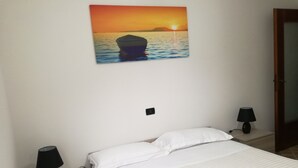 A bedroom with a bed, two bedside lamps, a nightstand, and a wall-mounted picture of a sunset over water.