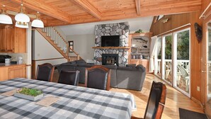 Interior acogedor de una cabaña con comedor, chimenea de piedra, televisor y una escalera que conduce a un balcón.