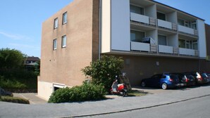 Ein mehrstöckiges Gebäude mit Ziegelaußenwand und weißen Obergeschossen. Ein Parkplatz mit mehreren Autos und einem roten Roller ist vorhanden.