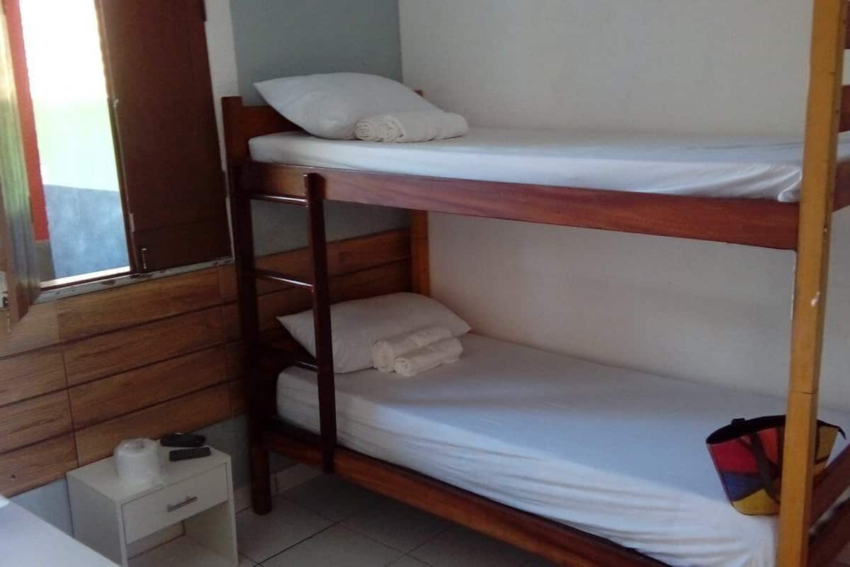 Una cama litera con ropa blanca y estructura de madera. Se ve un pequeño mesitas de noche blancas con un control remoto y una botella. Se observa una ventana con cortina.