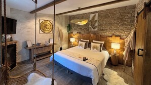 Un dormitorio con una cama grande, cabecera de madera, sillón colgante, pared de piedra y televisor montado.
