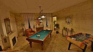 A room with a pool table, a foosball table, a fireplace, a leather armchair, and a chandelier.