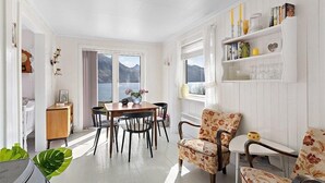 A bright room with a wooden dining table and chairs, a floral armchair, and a bookshelf with books and decorative items. - Private Cozy House by the Fjord (Flakstad)