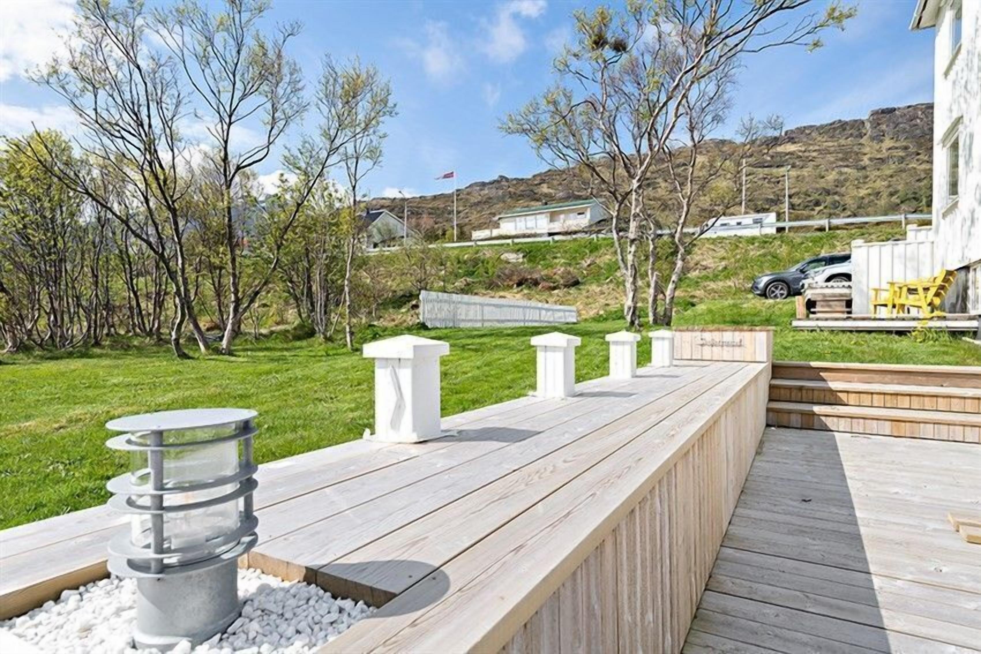Un terrazzo in legno con barbecue integrato e zona salotto, circondato da alberi e un pendio sullo sfondo.