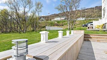 Un terrazzo in legno con barbecue integrato e zona salotto, circondato da alberi e un pendio sullo sfondo.