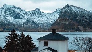 A white building with a dark roof, situated by a body of water with snow-capped mountains in the background. - Private Cozy House by the Fjord (Flakstad)