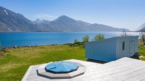 A wooden deck with a hot tub, a small blue shed, and a view of a lake with mountains in the background. - Private Cozy House by the Fjord (Flakstad)