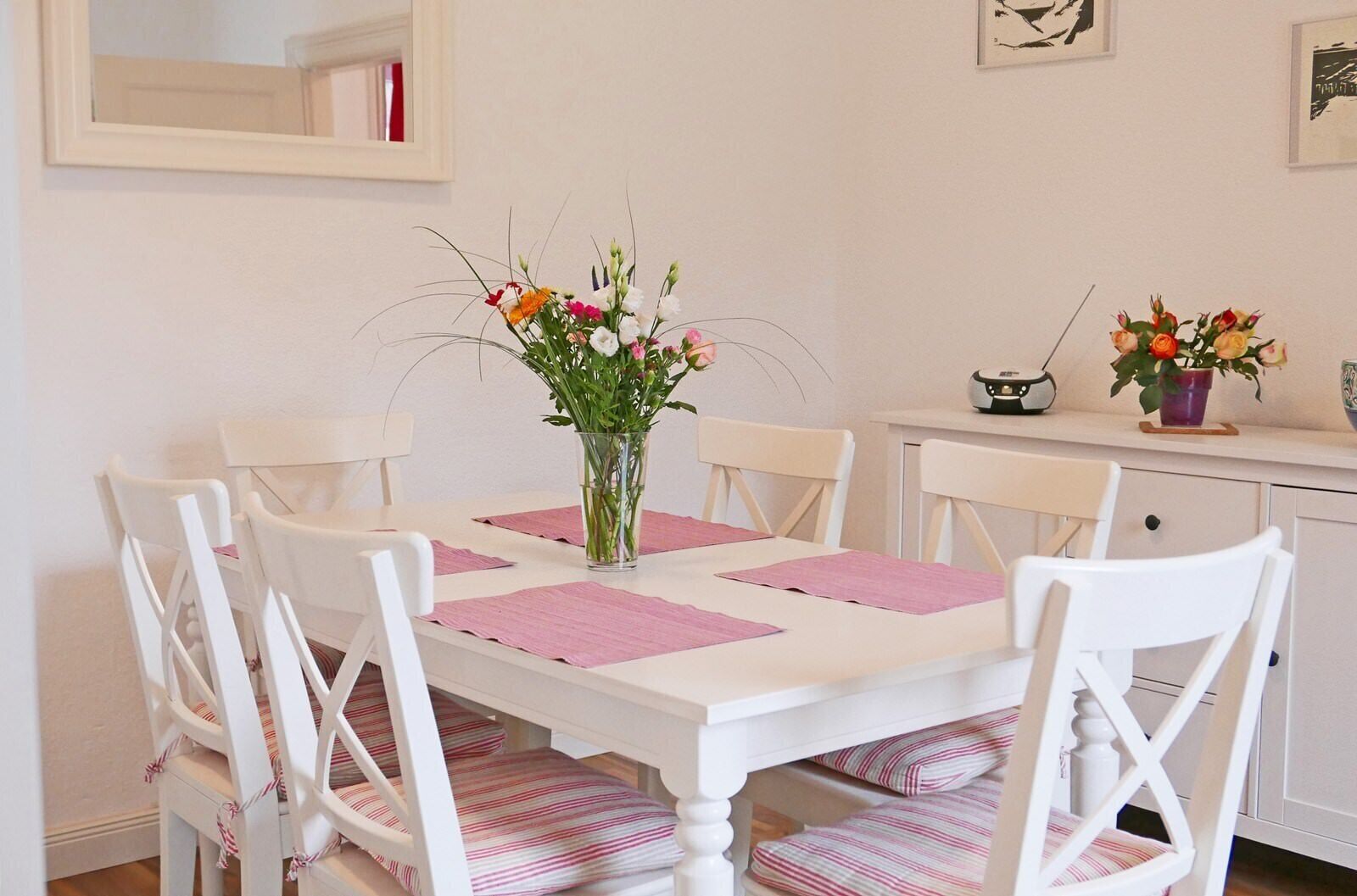 Uma mesa de jantar com seis cadeiras brancas, jogos americanos cor-de-rosa e um vaso de vidro transparente com flores.