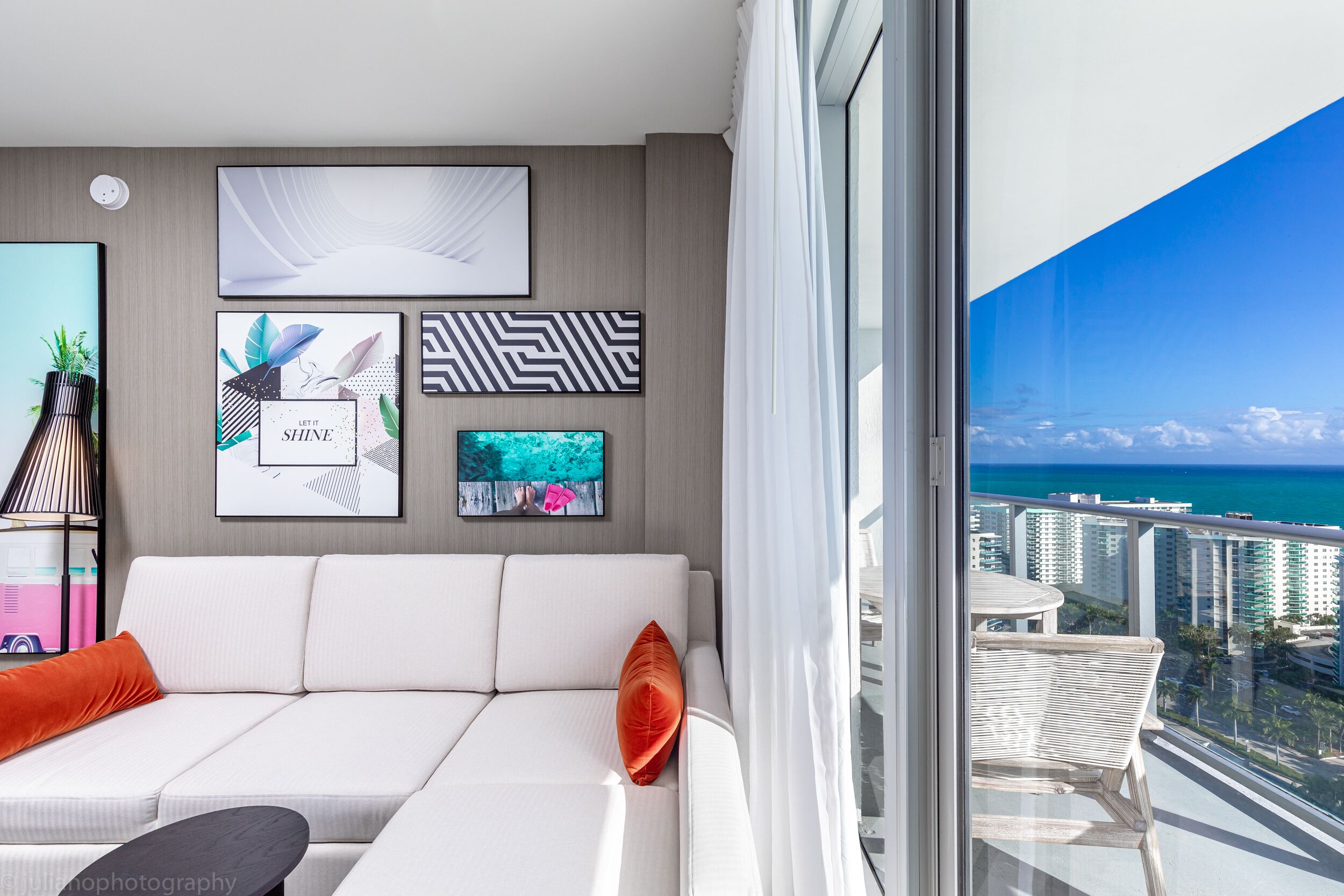 En moderne stue med en hvid sofa, et lille bord og en balkon med udsigt over havet og bygninger.