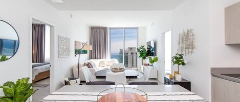 A modern, well-lit living room with a sofa, coffee table, and a dining area with a wooden table.