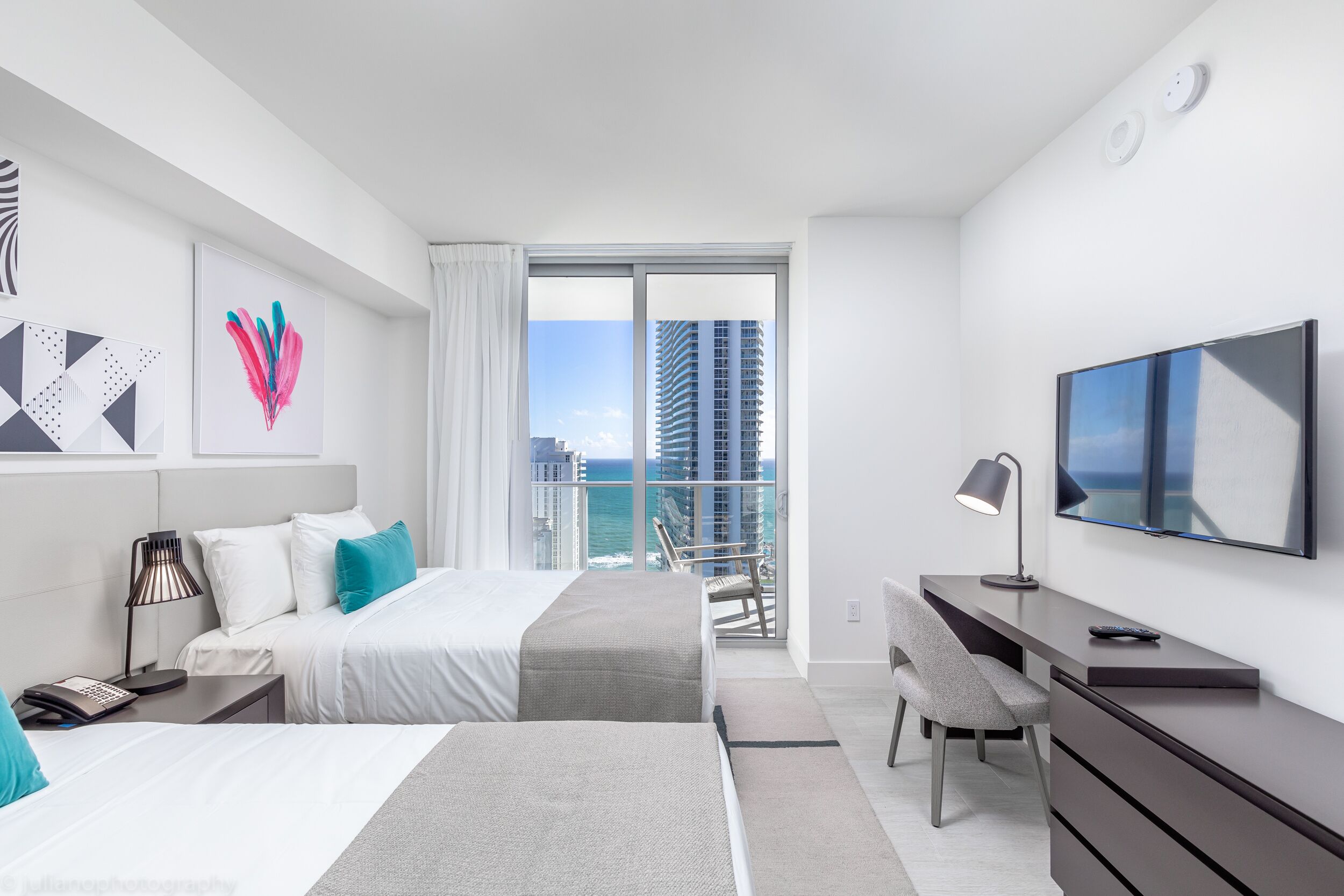 A modern hotel room with a balcony, a bed, a desk, a chair, and a TV.