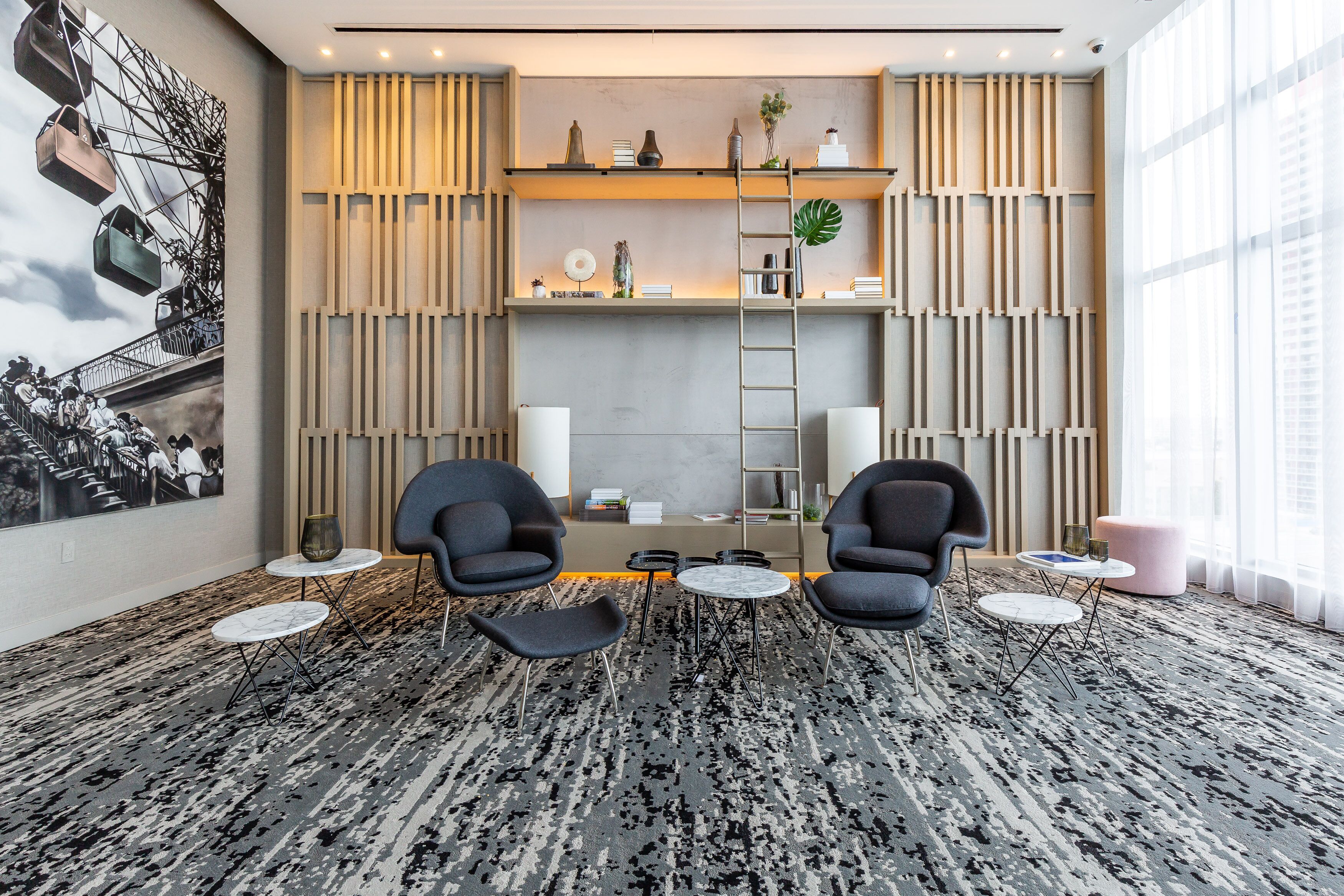 A modern hotel lobby with a large mural, two armchairs, a coffee table, and a bookshelf.
