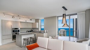 Une cuisine moderne dotée d’appareils électroménagers intégrés, un coin repas avec une table et des chaises, et une grande fenêtre offrant une vue sur l’océan et les immeubles de grande hauteur.