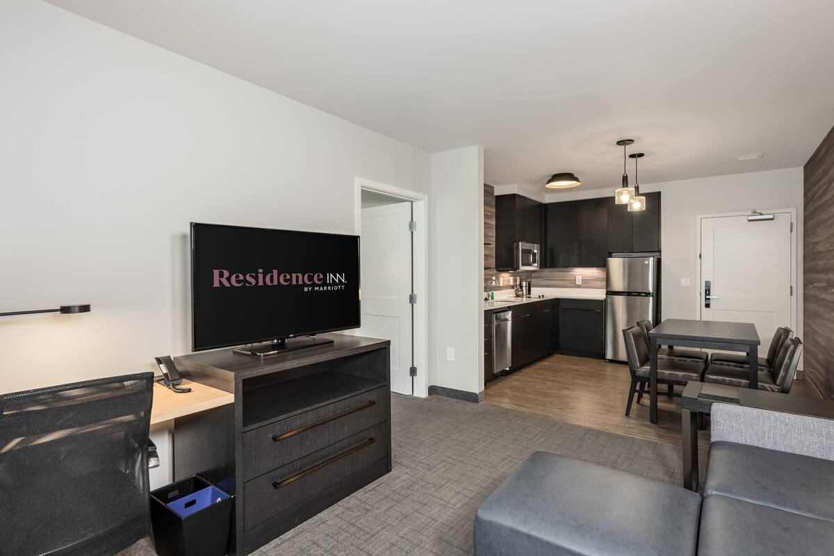 Een moderne woonkamer met een flatscreen televisie, een bank, een eettafel en een keukenhoek met donkere kastjes en roestvrijstalen apparatuur.