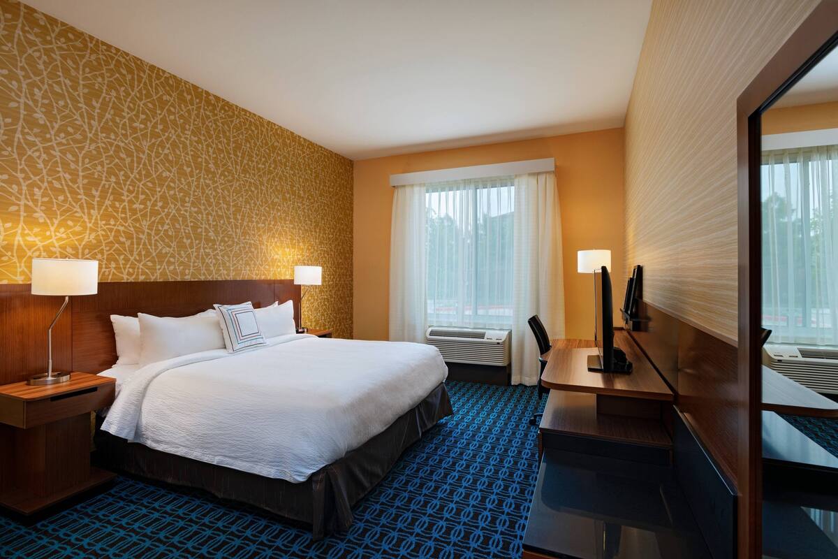 Una camera d'albergo con un letto grande, due comodini con lampade, una scrivania con una sedia, uno specchio ampio e una finestra con tende.