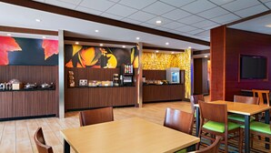A modern restaurant with wooden tables and chairs, a counter with a digital menu display, and a colorful mural on the wall.
