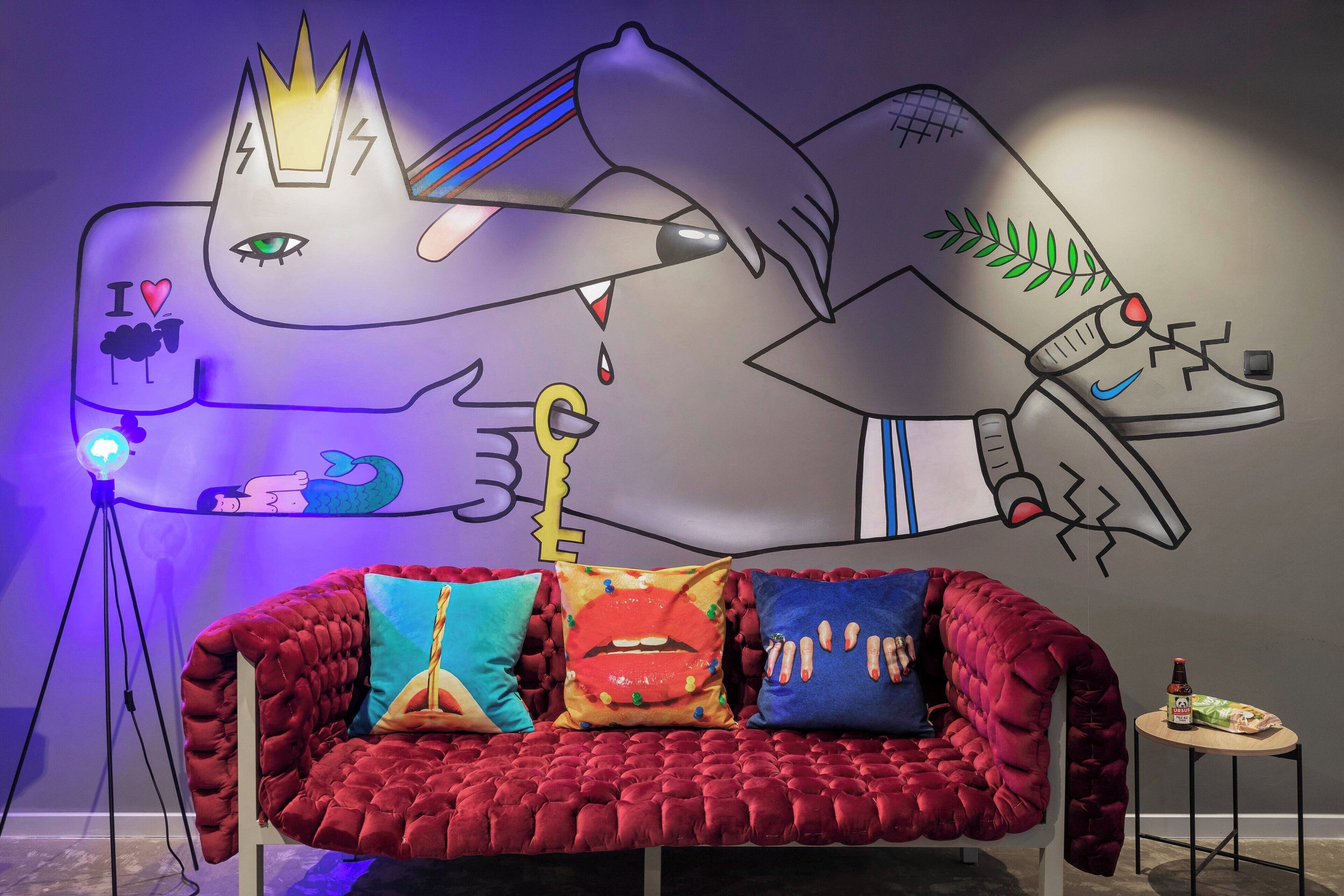 Une pièce avec un canapé en velours rouge, des coussins colorés, une fresque murale, un lampadaire sur trépied et une petite table sur laquelle se trouve une bouteille.
