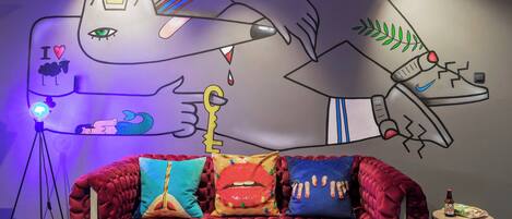 Una habitación con un sofá de terciopelo rojo, cojines coloridos, un mural en la pared, una lámpara de trípode y una pequeña mesa con una botella.