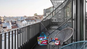 Un balcón con vista a la ciudad, una silla de metal, una mesa con una bebida y un bolso colorido.