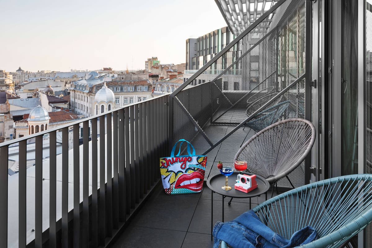 A balcony with a view of the city, a metal chair, a table with a drink, and a colorful bag.