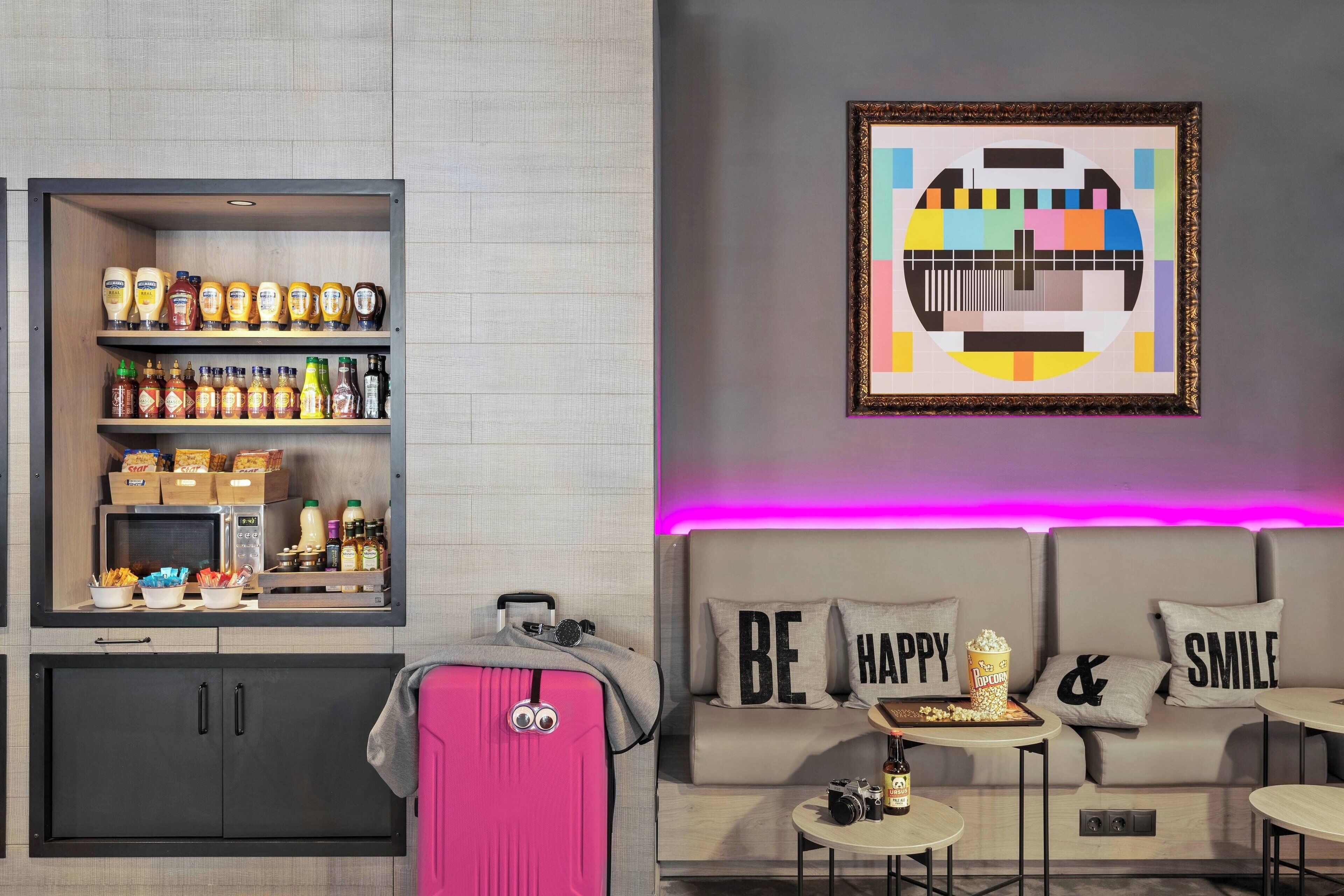 Une chambre d’hôtel moderne équipée d’une kitchenette, d’une valise rose, d’un canapé agrémenté de coussins aux motifs inspirants et d’une œuvre d’art accrochée au mur.