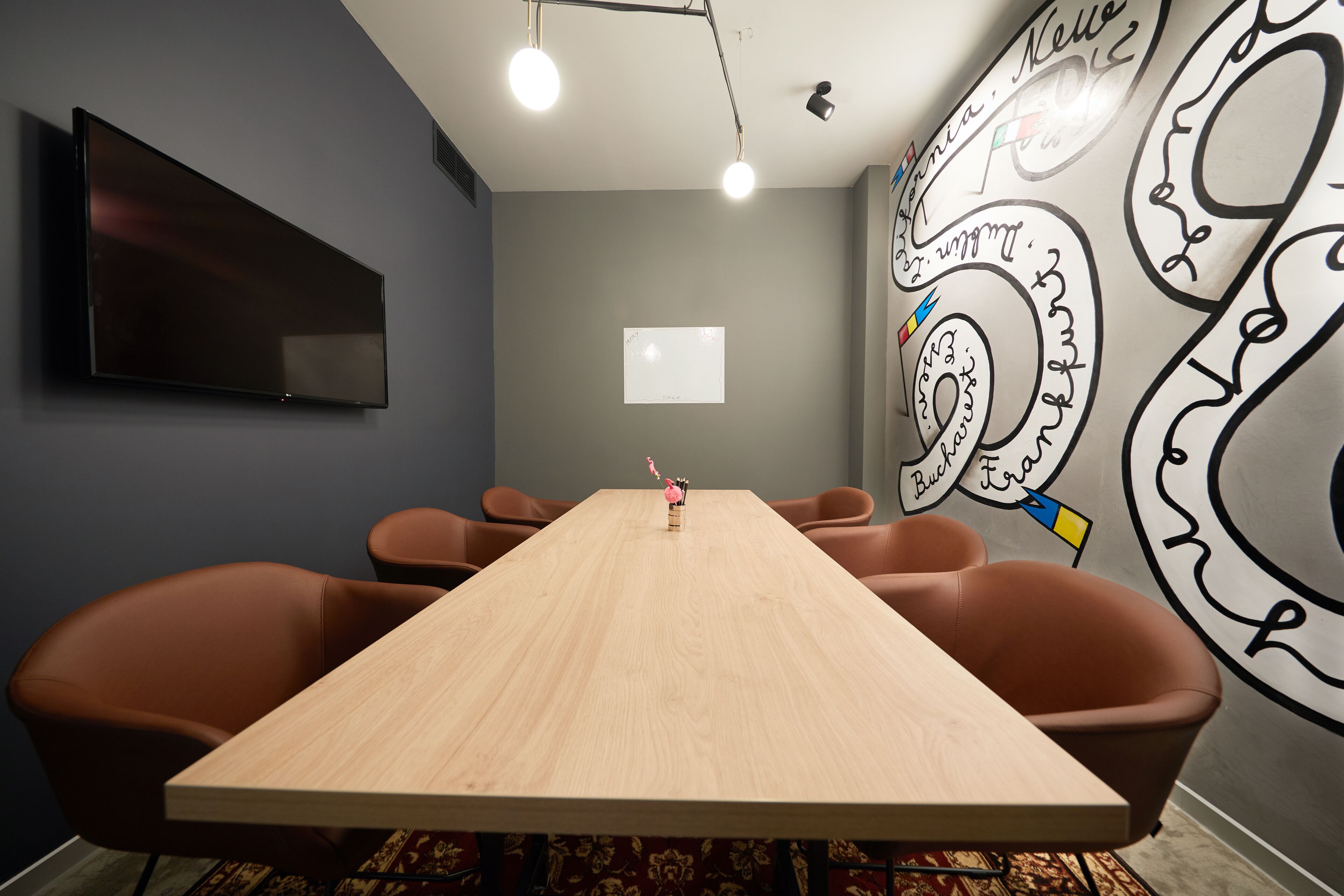 Une salle de conférence avec une longue table, des chaises marron, un téléviseur à écran plat, un écran de projection et un mur orné de dessins abstraits.