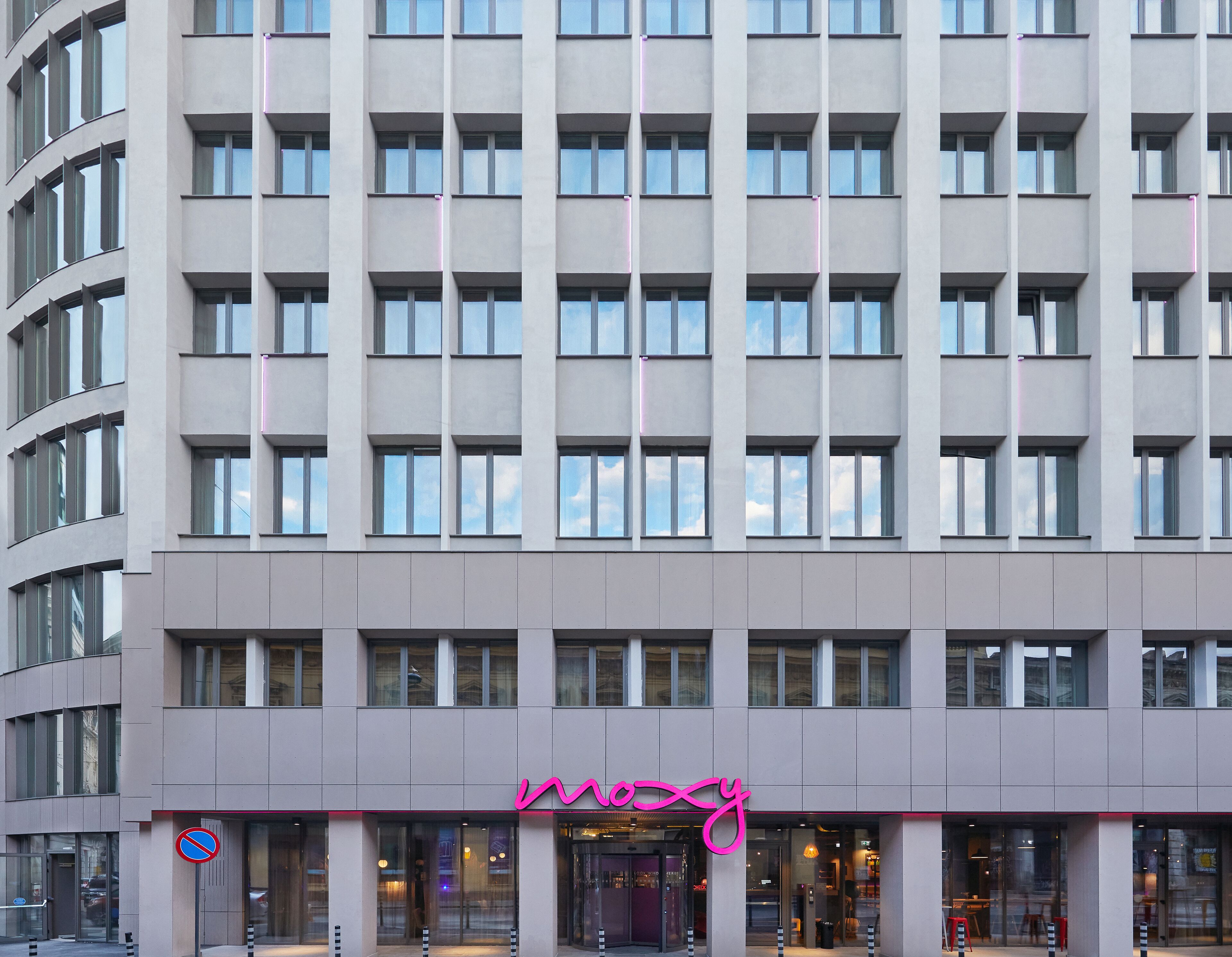 A multi-story hotel with a modern facade, featuring a prominent signage with a pink neon outline.
