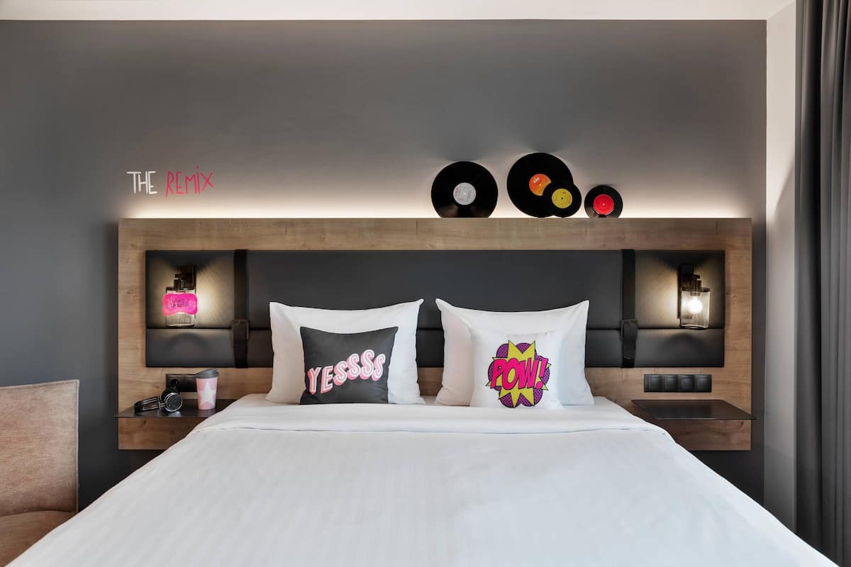 A modern hotel room with a wooden headboard, two decorative pillows, a bedside table with a lamp, and a wall-mounted shelf with vinyl records.