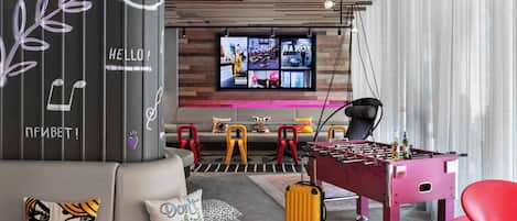 A modern hotel room with a foosball table, a sofa with decorative pillows, a television displaying multiple channels, and a wooden ceiling with pendant lighting.