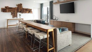 A modern living space with a wooden dining table, bar stools, a sofa, and a TV mounted on the wall. - Element by Marriott Iowa City (Iowa City)