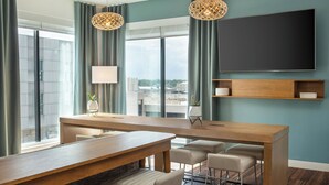 A modern hotel room with a wooden dining table, bar stools, a flat-screen TV mounted on the wall, and large windows with curtains. - Element by Marriott Iowa City (Iowa City)