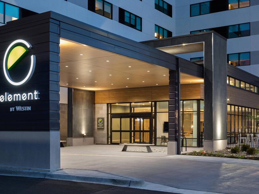 En moderne hotelindgang med et tydeligt logo, et overdækket udeareal og en veloplyst facade.