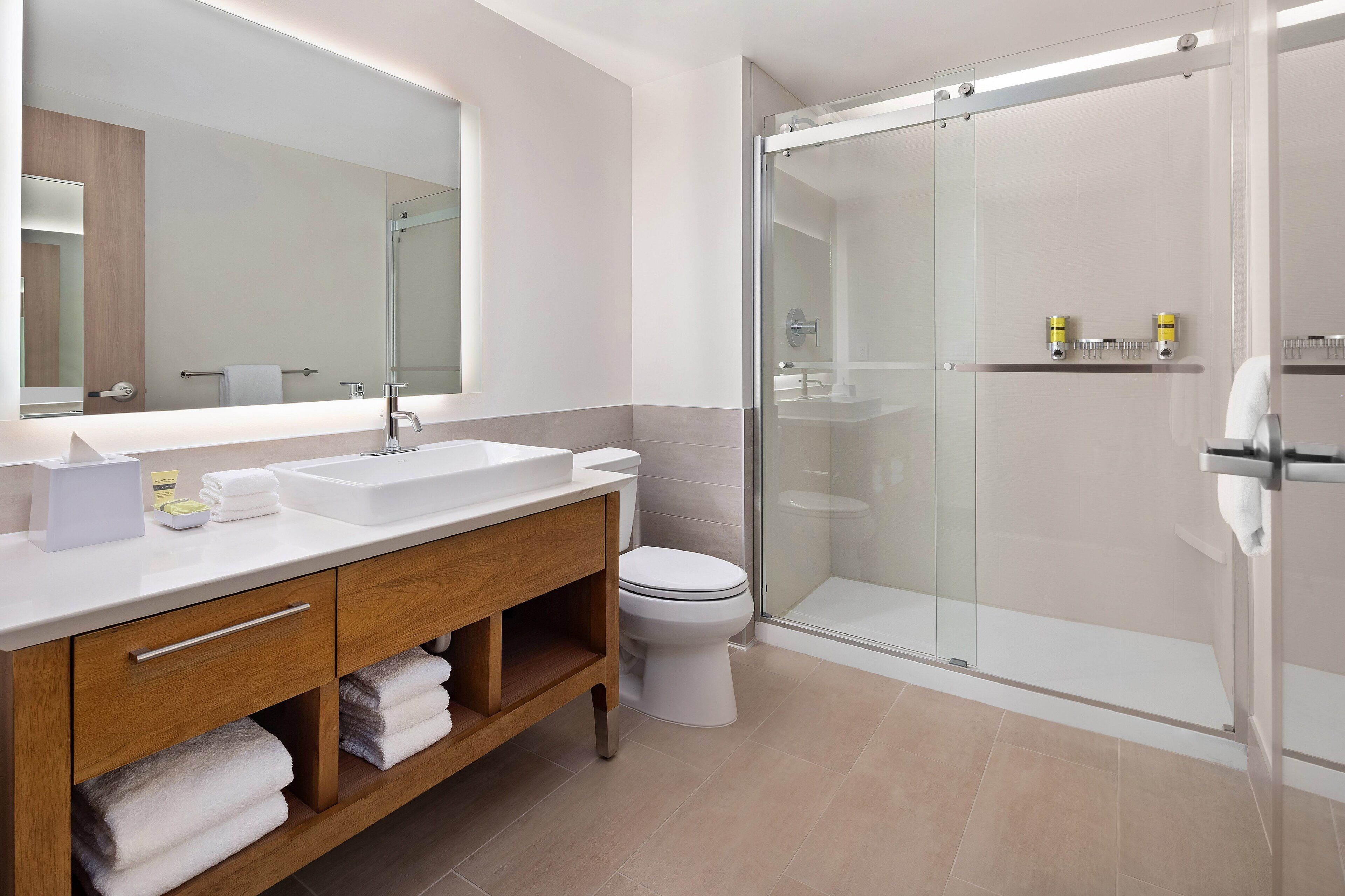 Une salle de bain moderne dotée d’une douche à parois de verre, d’un grand miroir et d’un meuble-lavabo en bois avec un espace pour le lavabo et des étagères pour les serviettes.