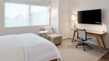 A hotel room with a bed, a desk with a chair, a TV, a window with a view, and a lamp.