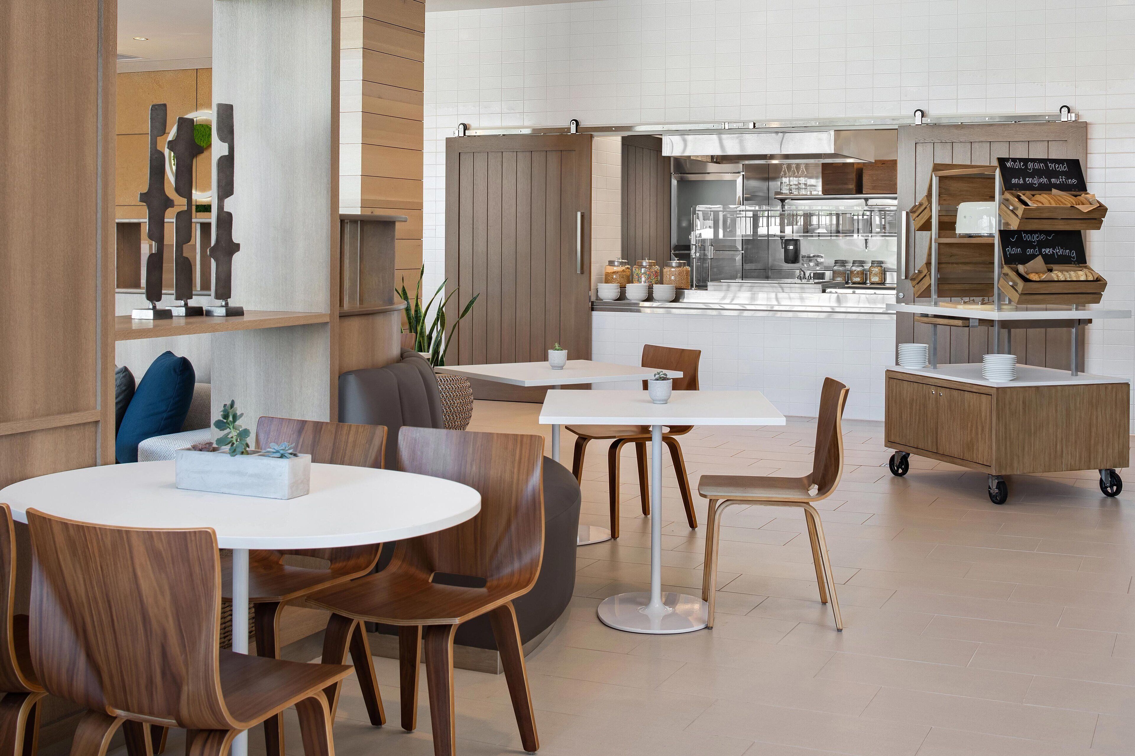 A modern cafe with round tables and wooden chairs, a kitchen area in the background, and a wooden counter with jars of bread and pastries.