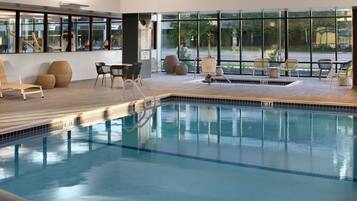 Una piscina interior con paredes de vidrio transparente, suelo de madera y varias zonas de estar con sillas y mesas.