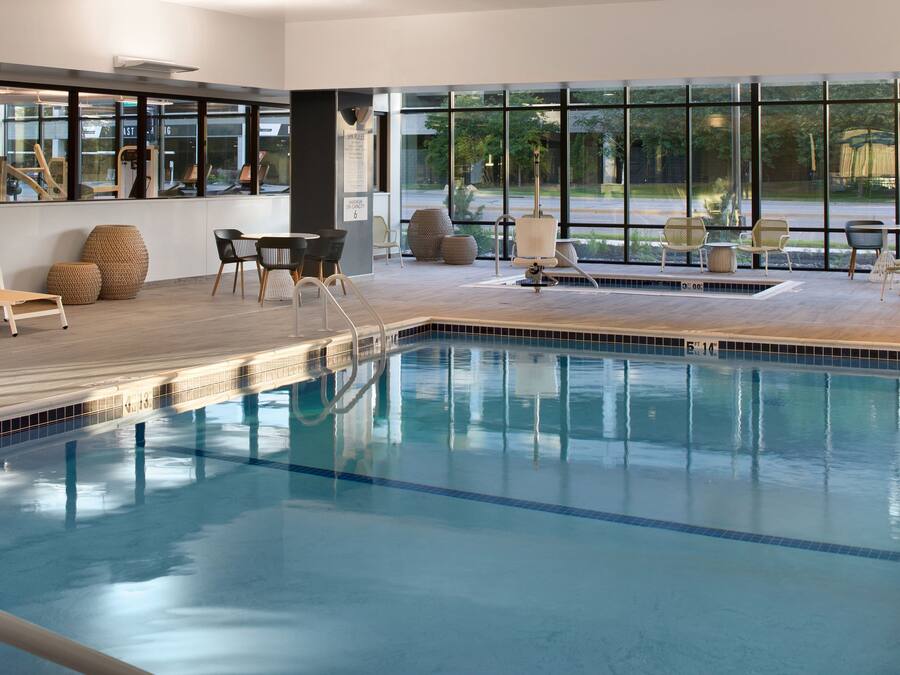 Una piscina interior con paredes de vidrio transparente, suelo de madera y varias zonas de estar con sillas y mesas.