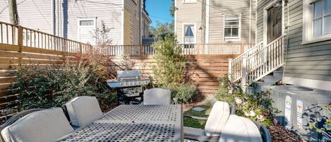 A patio with a metal table and white chairs, surrounded by a wooden fence and a small garden with flowers and shrubs.