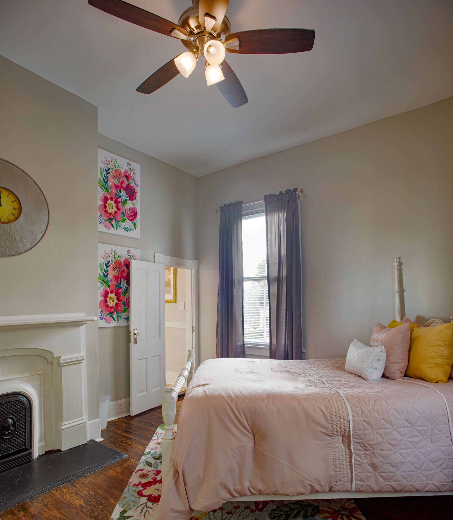 A bedroom with a bed, a ceiling fan, a fireplace, and floral artwork on the walls.