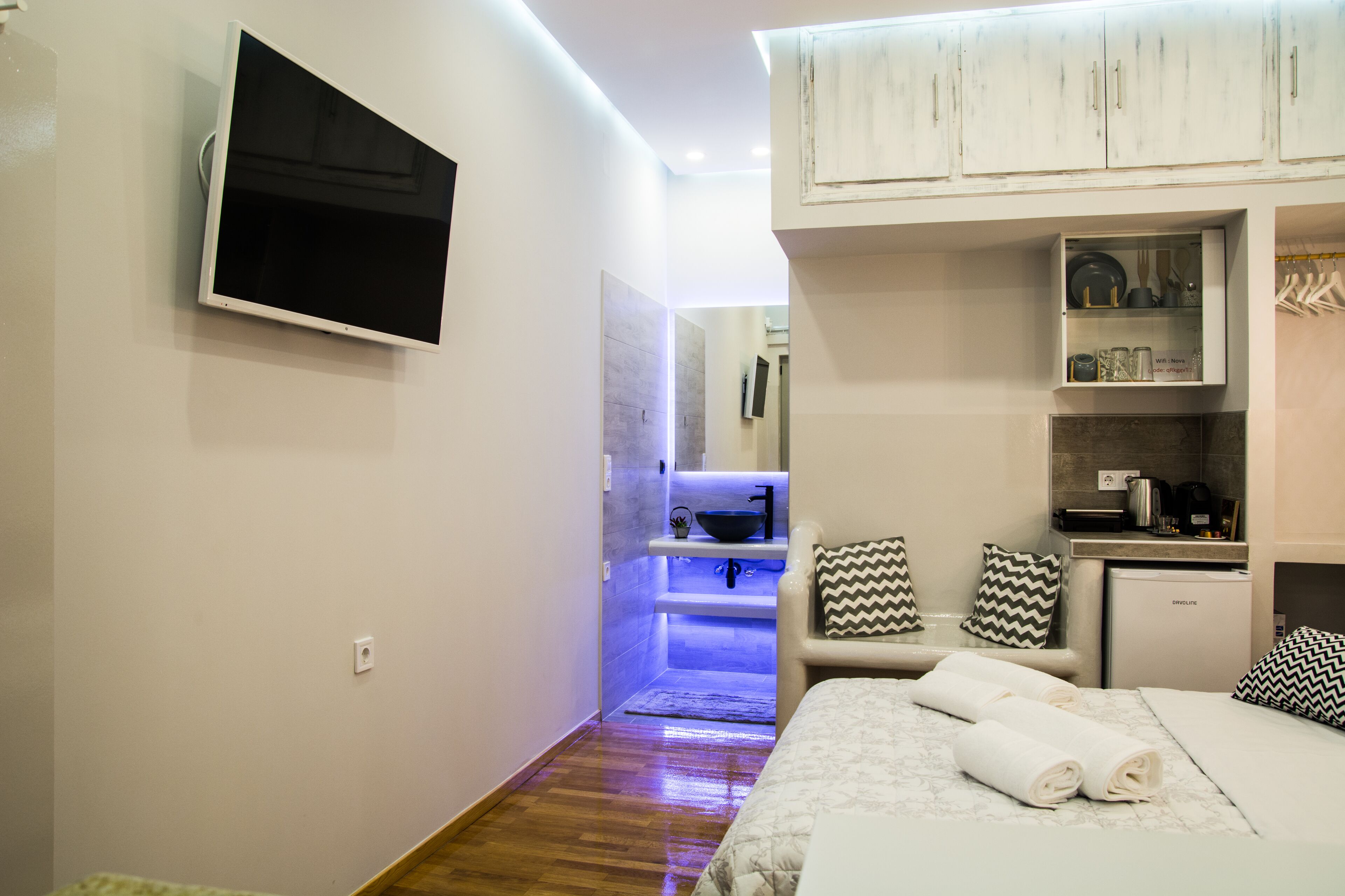 Une chambre d’hôtel comprenant un lit, un téléviseur fixé au mur, un coin cuisine et une salle de bain avec un lavabo éclairé de lumière bleue.