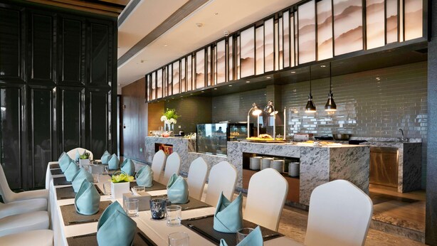 A modern dining area with a long table, white chairs, and a kitchen in the background. - Days Hotel Suites By Wyndham Zixin Changsha (Changsha)