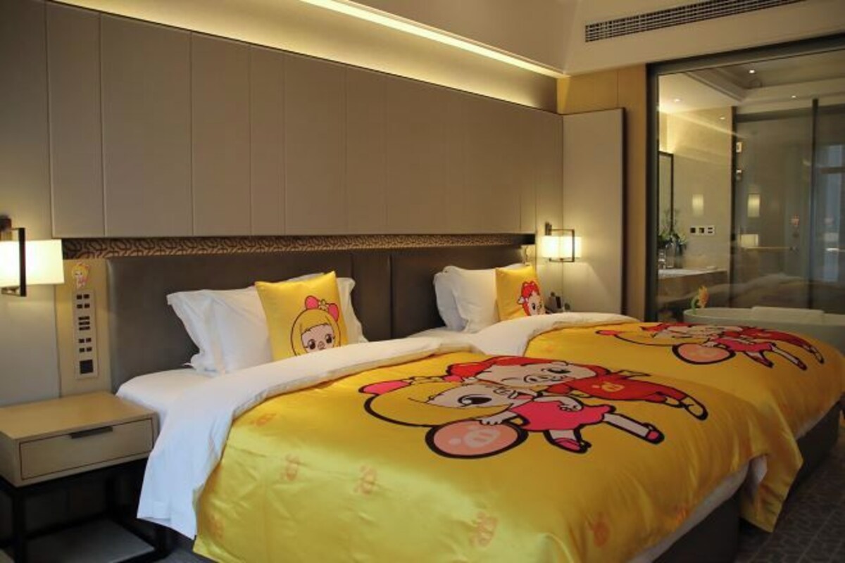 Une chambre d’hôtel avec deux lits, chacun recouvert d’une couverture jaune ornée de personnages de dessins animés, une table de chevet avec une lampe et une grande fenêtre donnant sur la ville.